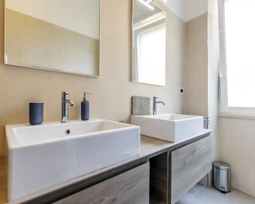 Modern double-sink bathroom with rectangular vessel sinks, wall-mounted mirrors and natural light from a large window. Clean, contemporary finishes with light beige tiles and wood-effect vanity.