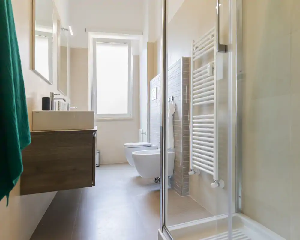 Modern, bright bathroom with a shower enclosure, wall-hung toilet and bidet, vanity with sink, and a towel radiator. Neutral tones and a large frosted window provide natural light.