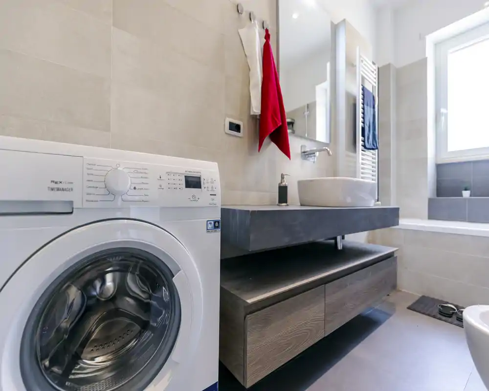 Modern bathroom with a front-loading washing machine, floating vanity with vessel sink, mirror and bright window providing natural light.