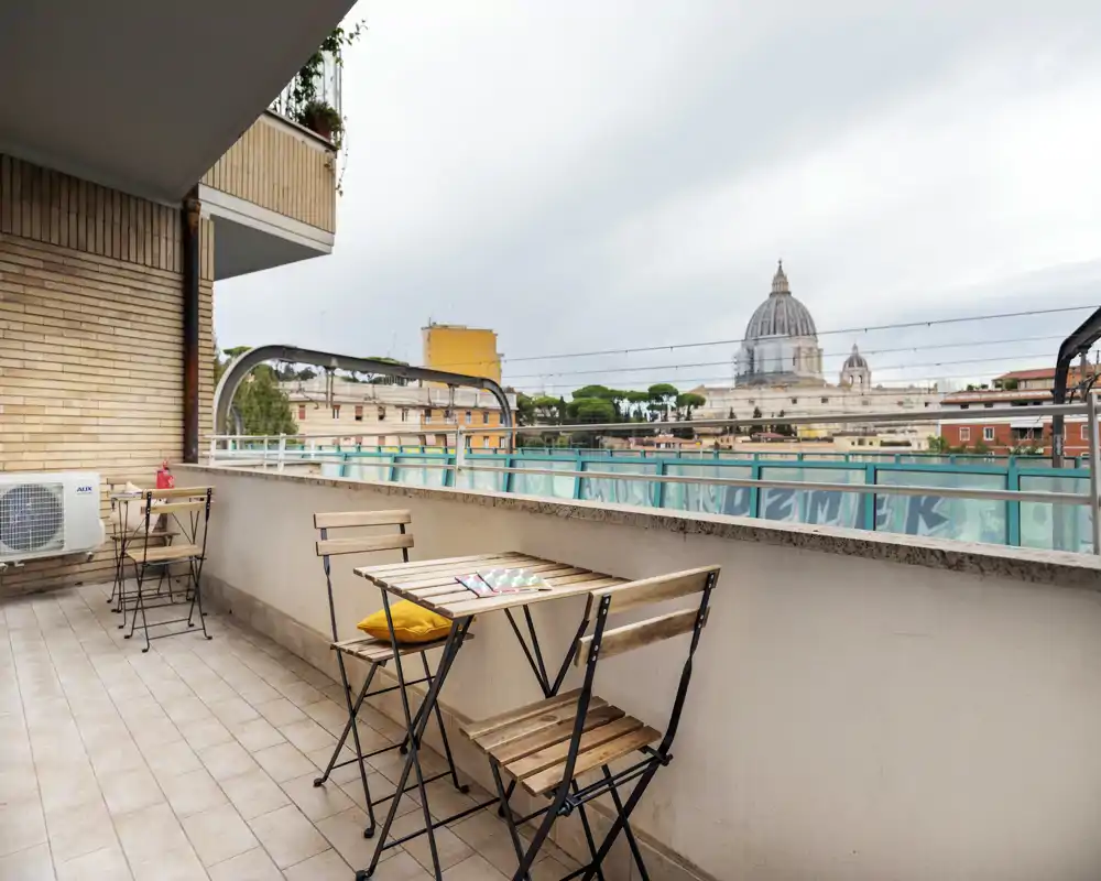 Sunny furnished balcony with a small wooden bistro table and folding chairs, overlooking a city skyline with a domed landmark — ideal for morning coffee.