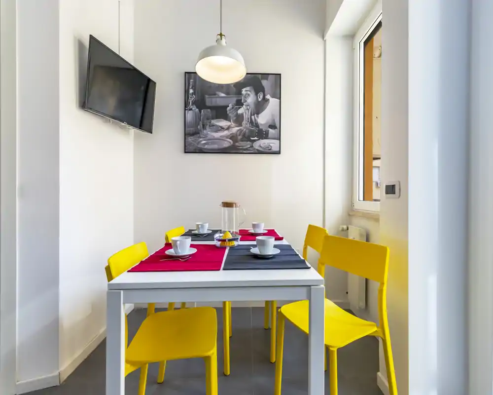 Bright, modern dining corner with a white table set for four and vivid yellow chairs, lit by a pendant lamp and natural light from the window.