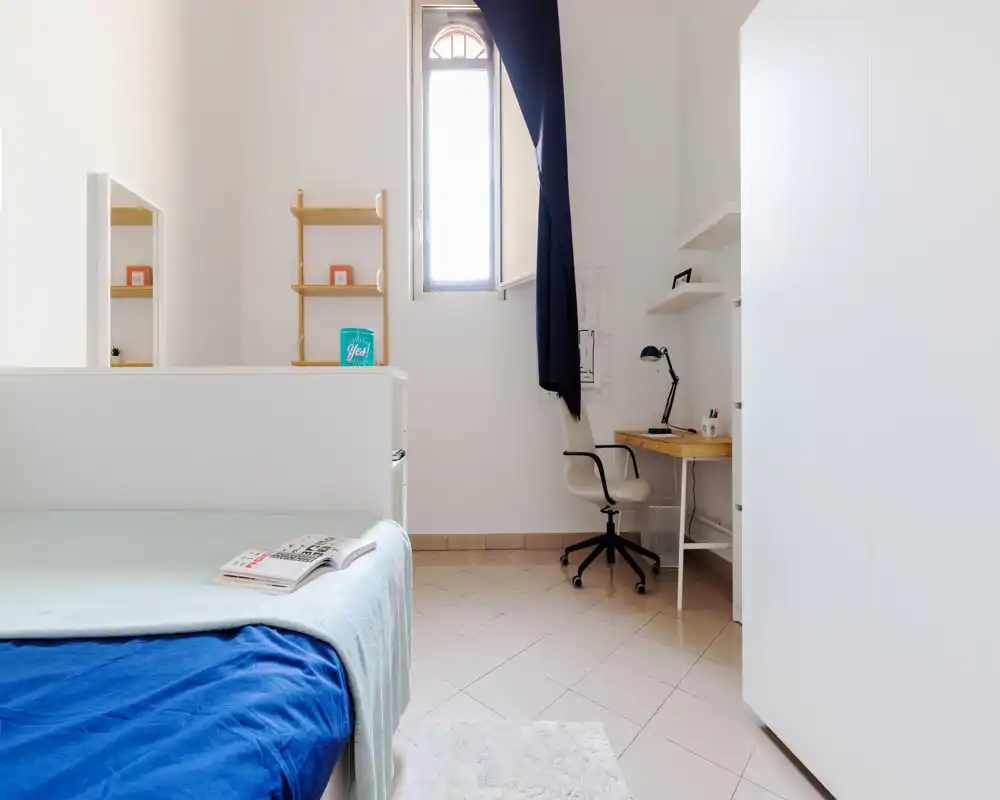 Bright, minimal bedroom with a single bed in the foreground, a tidy desk and shelving by a tall window—clean, airy and well lit for study or rest.