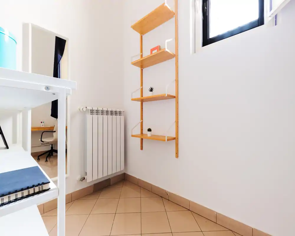Small bright entrance or hallway corner with tiled floor, wall-mounted radiator and minimalist wooden shelving — clean, simple transition space.