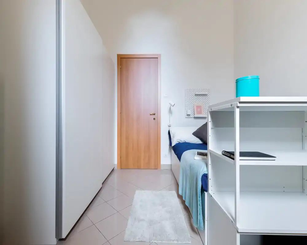 Neat single bedroom with a clean modern wardrobe, a well-made bed with pillows and a cozy throw, plus shelving for storage — bright, minimal and ready for a guest.