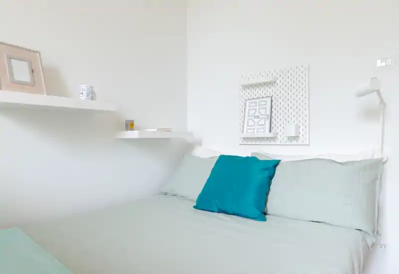 Bright, minimal bedroom with a neatly made double bed, teal accent cushion and simple wall shelves—ideal for a calm, modern stay.