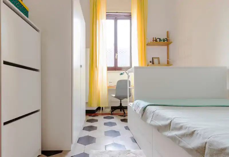 Bright, tidy bedroom with a single bed in the foreground, a compact study desk by the window and warm yellow curtains creating a welcoming atmosphere.