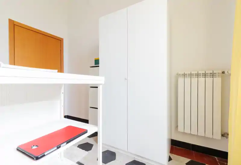 Minimal bedroom corner with a white wardrobe, radiator and a portion of a white desk; bright, clean and simply furnished.