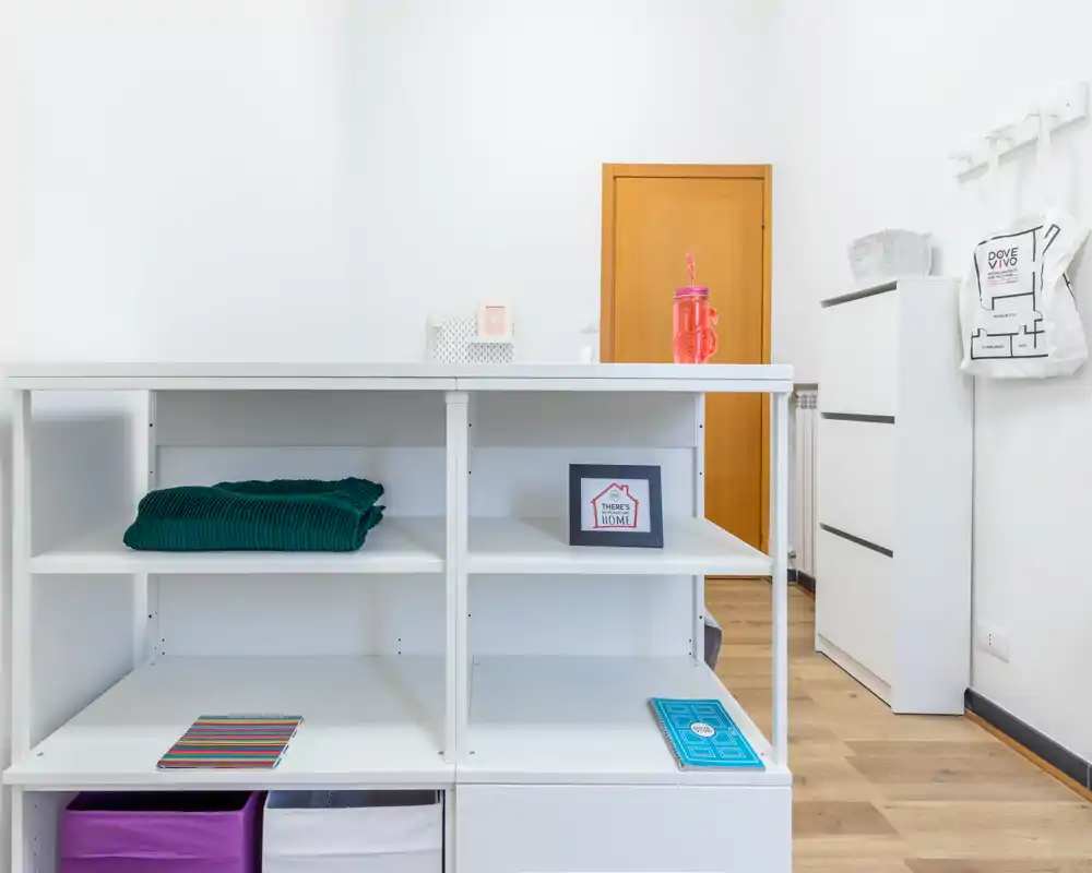 Ingresso luminoso con una scaffalatura bianca usata come contenitore/divisorio, pavimento in legno chiaro e una cassettiera semplice; ambiente pulito, minimale e funzionale per organizzare scarpe e piccoli oggetti.