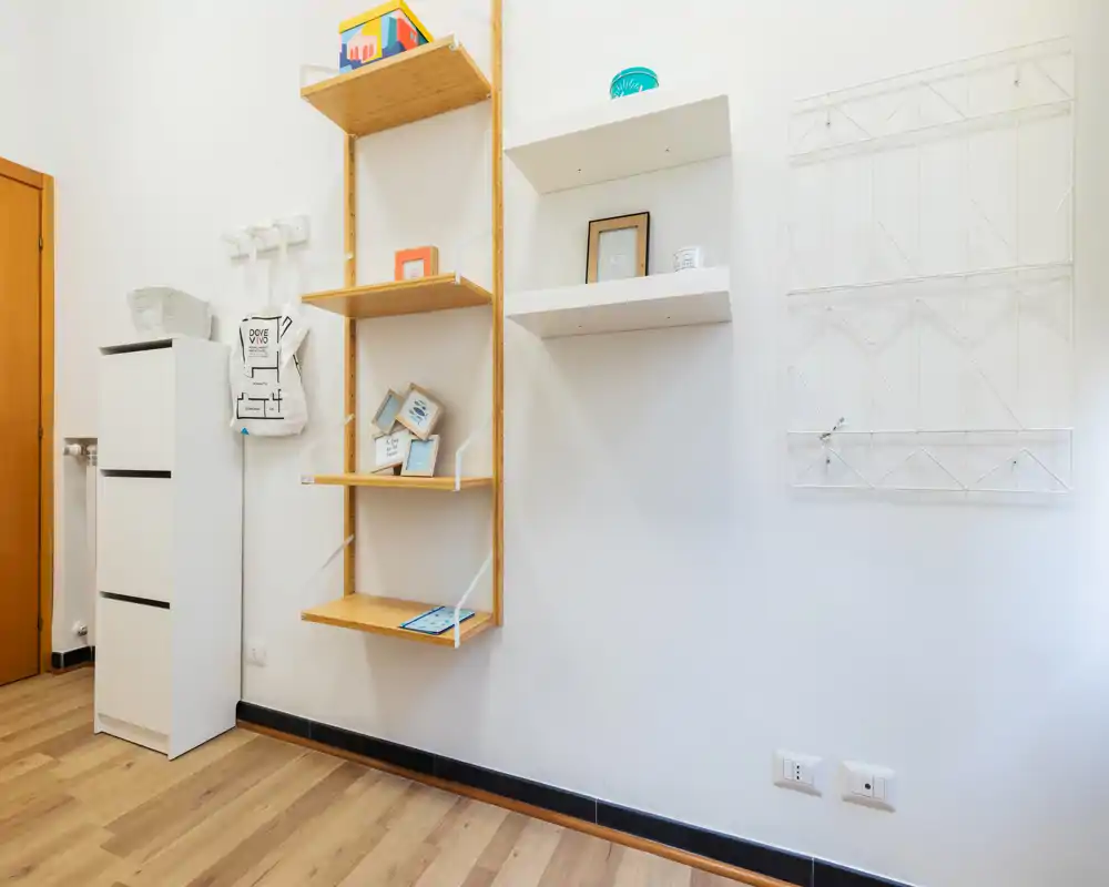 Angolo d'interno minimale con mensole in legno a parete, un armadietto bianco sottile, appendiabiti a muro e pavimento in legno chiaro. L'immagine mette in evidenza scaffalature e contenimento decorativo più che una funzione d'uso specifica.