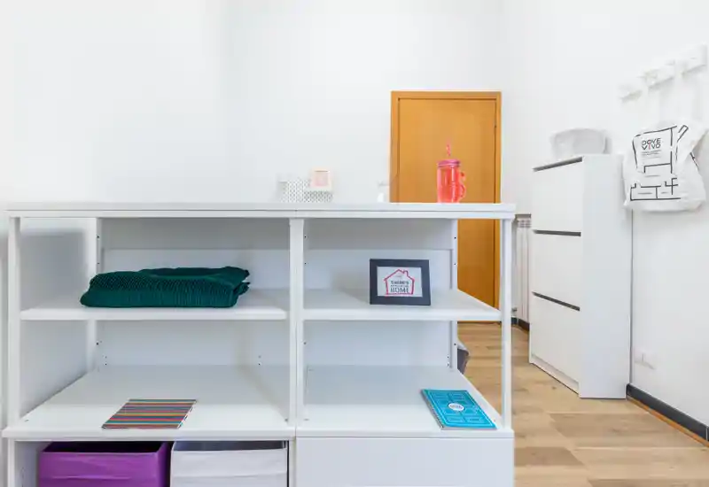 Area living luminosa e minimal con scaffalature aperte, dettagli decorativi e pavimento in legno: ambiente pulito e ben illuminato ideale per un soggiorno compatto o monolocale.