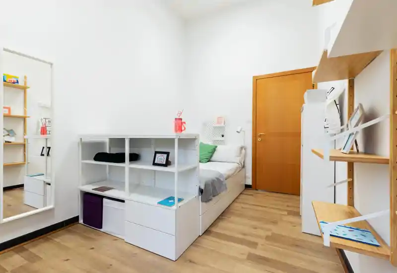 Camera da letto compatta e luminosa con letto singolo, scaffalatura integrata ai piedi del letto e mensole in legno minimaliste — pareti bianche e pavimento in legno che donano un ambiente pulito e accogliente.