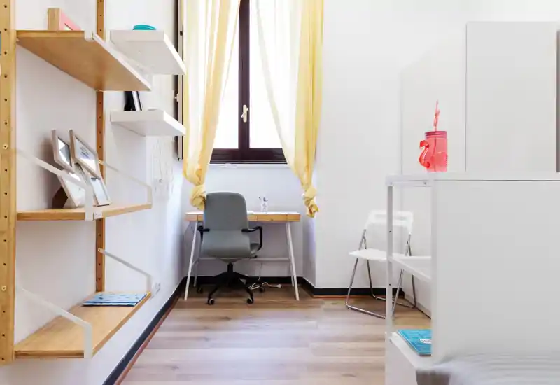 Angolo luminoso e ordinato con scrivania vicino alla finestra, scaffalatura in legno e sedute moderne — perfetto per un monolocale compatto.