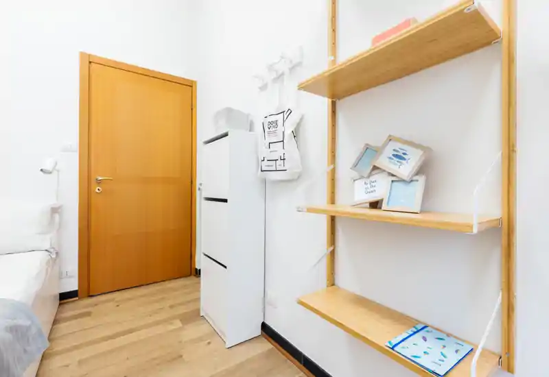 Angolo di camera da letto luminoso, con bordo del letto visibile, porta in legno, sottile mobiletto bianco e mensole in legno a parete con cornici.