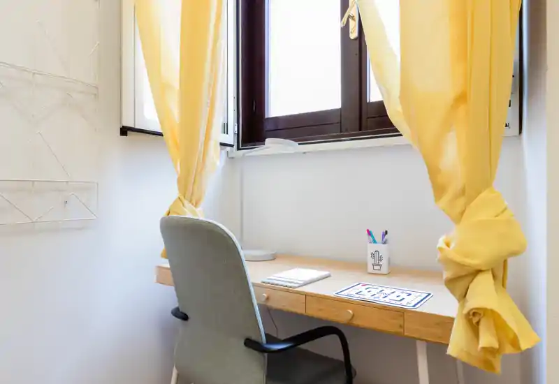 Un angolo studio luminoso e ordinato vicino alla finestra, con scrivania in legno, sedia comoda e tende gialle che creano un ambiente accogliente per lavorare o studiare.