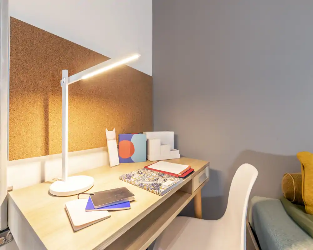 Rincón de dormitorio luminoso y cuidado con escritorio de madera y lámpara de trabajo junto a una cama parcialmente visible; papelería ordenada y cojines crean un espacio acogedor.