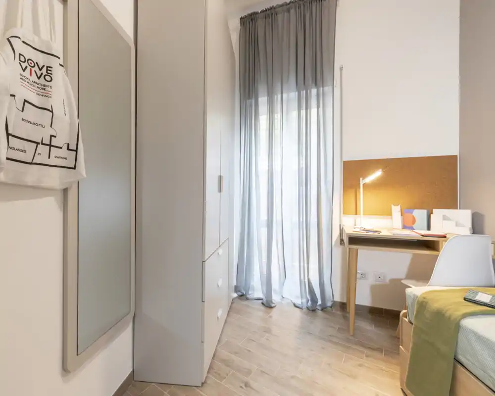 Rincón de dormitorio luminoso y minimalista con escritorio, armario y cama individual parcialmente visible; cortinas translúcidas aportan luz natural suave.