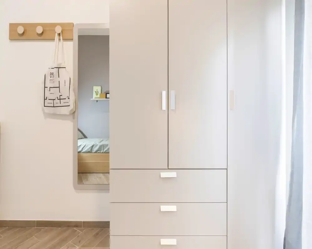 Dormitorio luminoso y minimalista, dominado por un armario y un pequeño vistazo de la cama a través de una abertura en la pared; líneas limpias y tonos neutros que transmiten calma y modernidad.