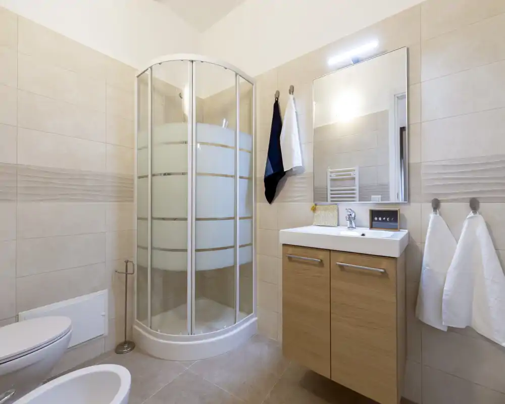 Baño moderno y compacto con ducha de esquina de vidrio, mueble lavabo suspendido con espejo y ganchos para toallas; azulejos neutros y buena iluminación que ofrecen un aspecto limpio.