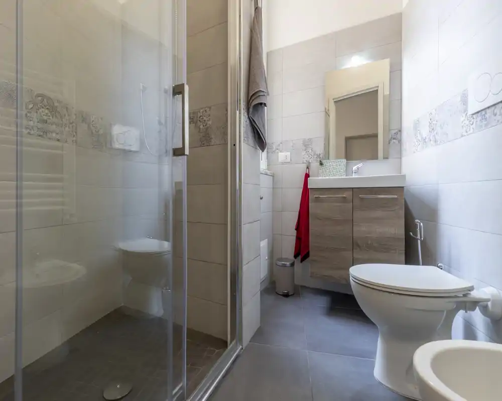 Baño moderno y bien iluminado con ducha de cristal, mueble con lavabo y espejo, inodoro y bidé; azulejos neutros y acabados cuidados.