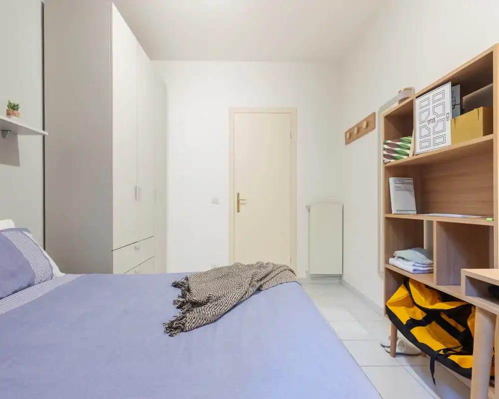 Camera da letto luminosa e moderna con letto ben sistemato in primo piano, armadio bianco e scaffalatura aperta per riporre oggetti. Tessili morbidi e palette neutra trasmettono un'atmosfera tranquilla e accogliente.