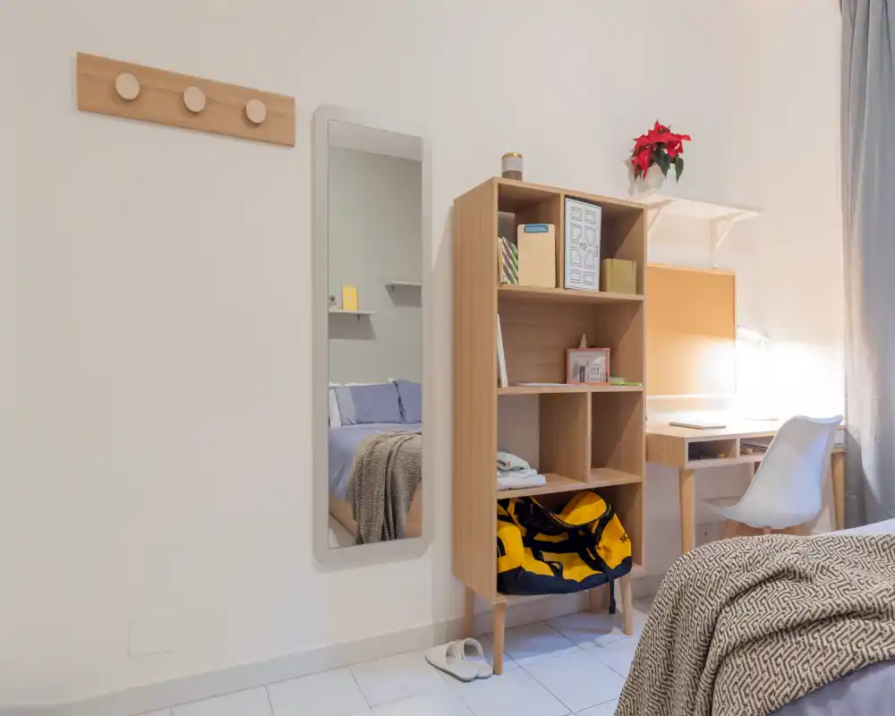 Angolo camera luminoso e ordinato con letto visibile, specchio a muro, scaffale e una piccola scrivania. Toni neutri e tessuti morbidi rendono lo spazio pratico e accogliente.