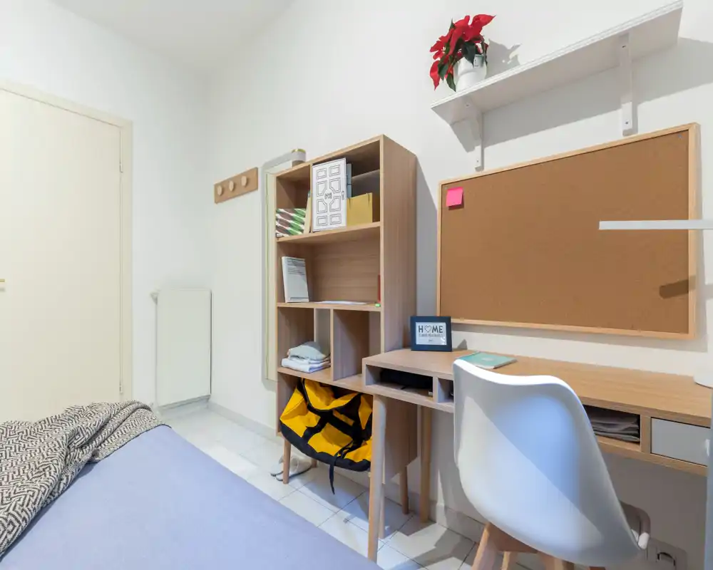Angolo camera compatto e luminoso con letto parzialmente visibile, libreria in legno e scrivania con bacheca — ideale per studenti o soggiorni brevi.