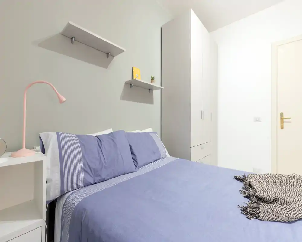 Camera da letto luminosa e ben curata con grande letto matrimoniale con biancheria azzurra, comodino con lampada e armadio bianco alto — ideale per annunci in affitto.