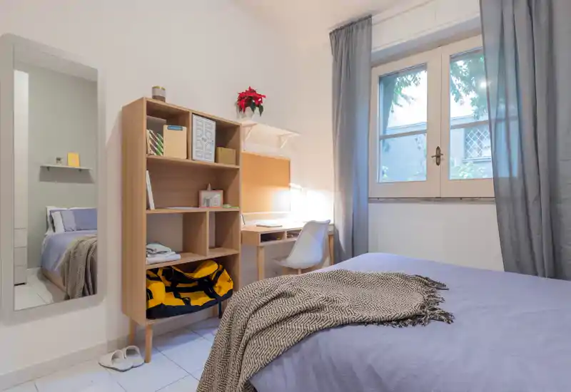 Camera da letto luminosa e ordinata con un letto coperto da una trapunta lilla, un accogliente plaid, una scrivania compatta con scaffali e una finestra che illumina la stanza.