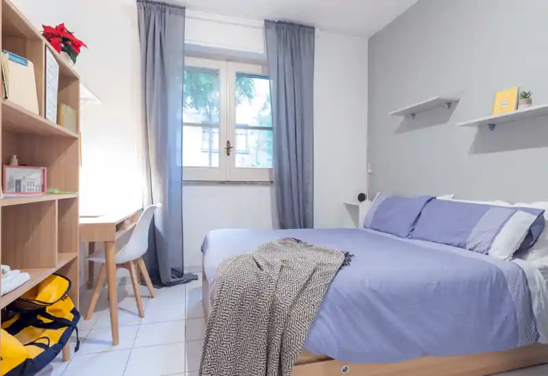 Camera da letto luminosa e ordinata con un letto matrimoniale con biancheria azzurra, un morbido plaid, mensola laterale e una scrivania vicino alla finestra che lascia entrare luce naturale.
