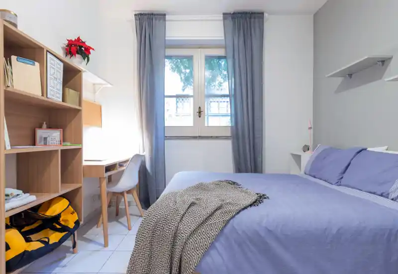 Camera da letto luminosa e ordinata con letto matrimoniale con biancheria blu, una coperta accogliente e una piccola scrivania vicino alla finestra — ideale per lavoro e riposo.