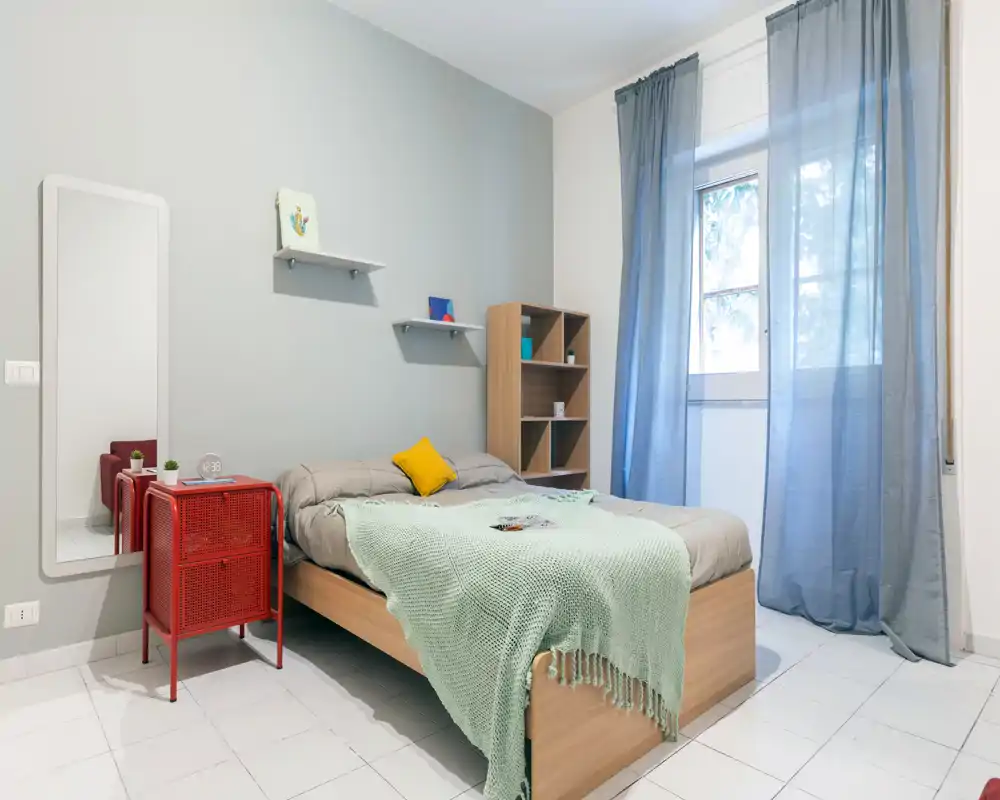 Bright, modern bedroom with a neatly made wooden bed, soft mint throw and a yellow accent cushion. The room features a tall window with sheer curtains, a red bedside cabinet and a full-length mirror, offering a fresh and inviting atmosphere.