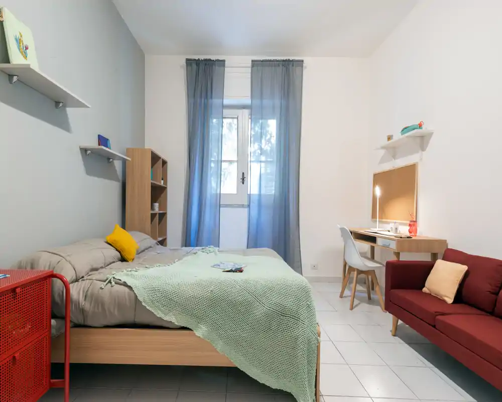Modern, well-lit bedroom with a double bed, soft green throw and a workspace by the window. The room features a small red sofa, shelving and a tidy desk ideal for remote work.