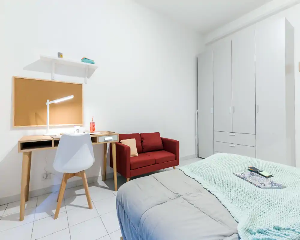 Bright, modern bedroom with a neatly made bed in the foreground, a compact wooden desk and chair, and a small red loveseat next to a large white wardrobe.