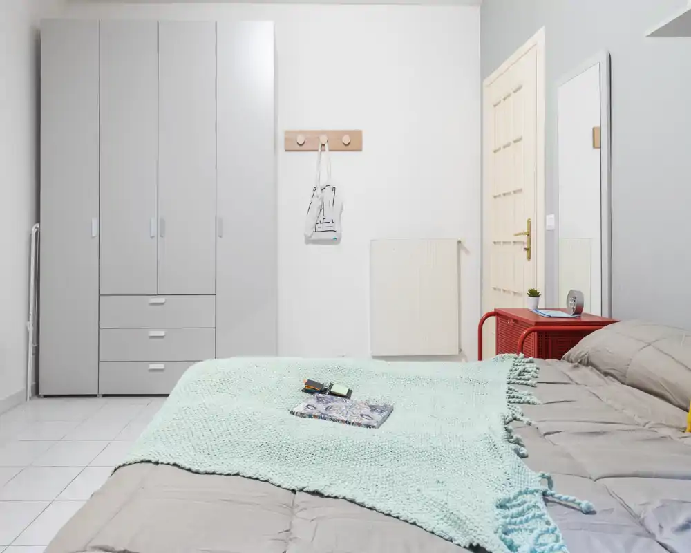 Bright, modern bedroom with a large bed covered by a soft mint throw, a wardrobe on the left and a bedside table with mirror on the right — clean, minimal and welcoming.