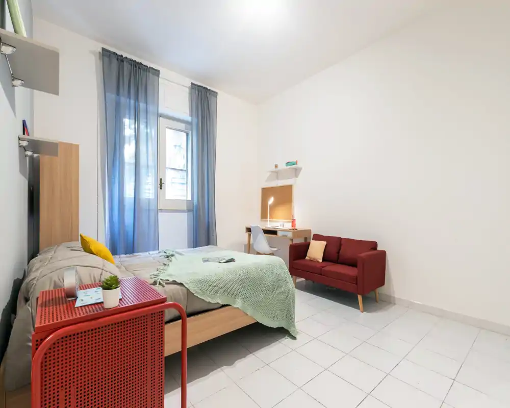 Bright, modern bedroom with a double bed, green throw and a small red sofa. Functional study corner and natural light from the window make this room ideal for comfortable stays.