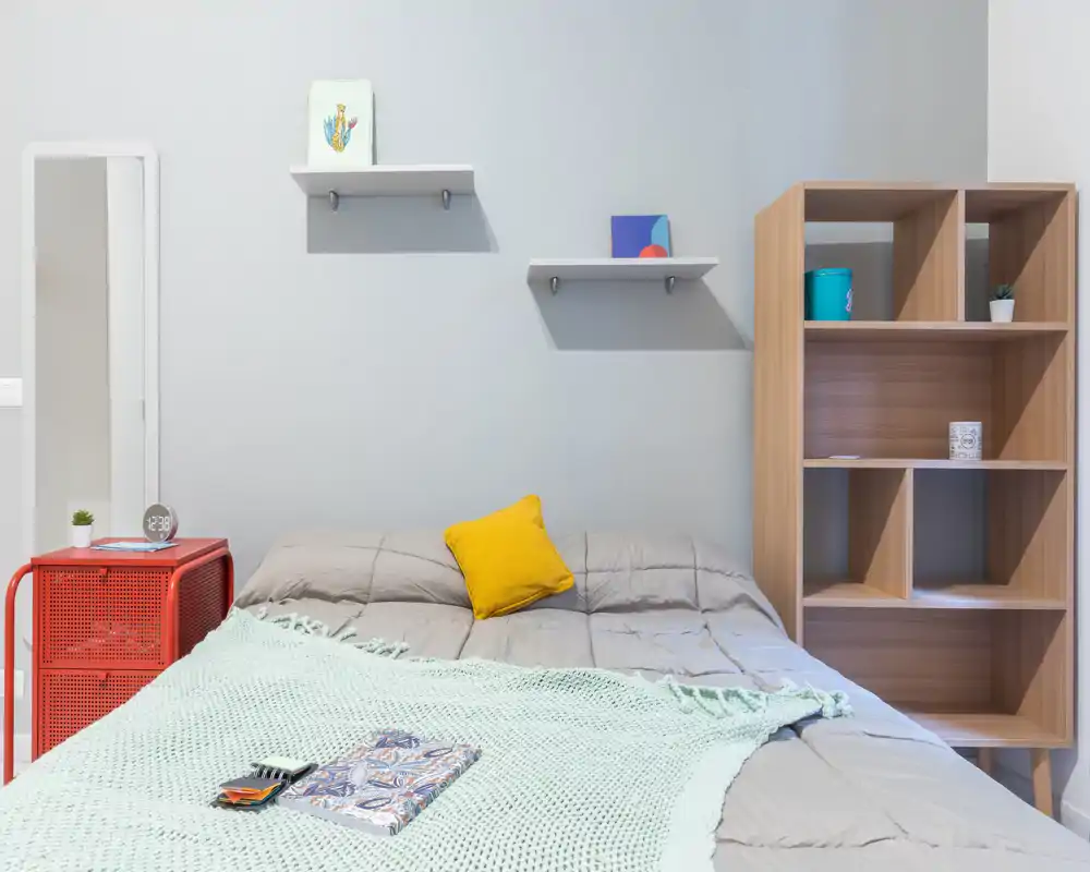 Bright, modern bedroom with a neatly made double bed, mint throw and a yellow accent cushion; shelving and a wooden cubby unit provide storage and personality.
