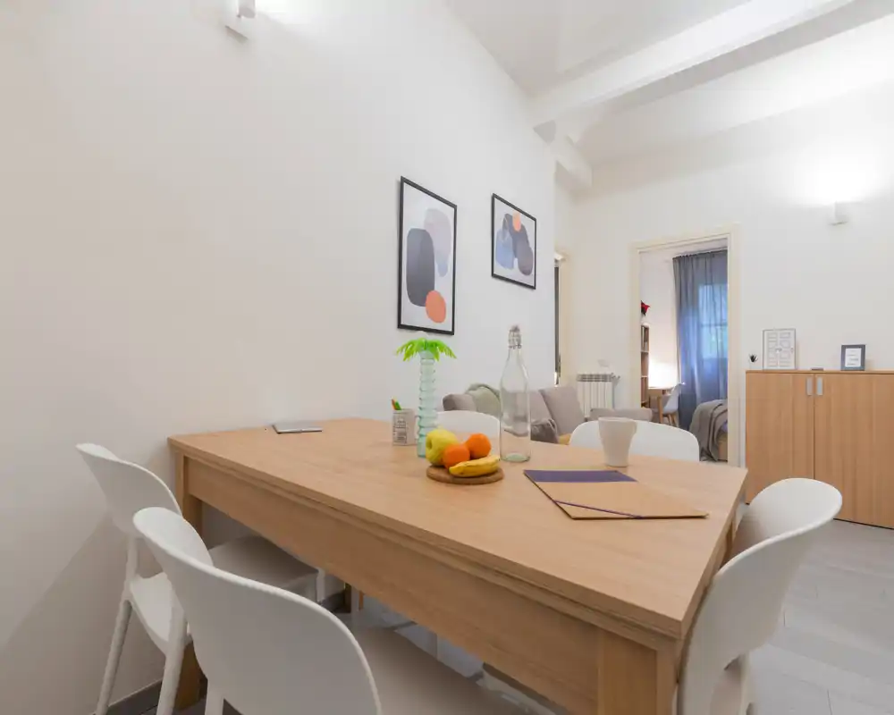 Zona pranzo luminosa e moderna con tavolo in legno, sedie bianche e stampe decorative; apparecchiata con una ciotola di frutta e una bottiglia per un'atmosfera accogliente.
