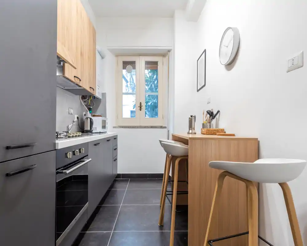 Cucina moderna compatta con basi scure, pensili in legno e un piccolo bancone con sgabelli. La luce naturale dalla finestra centrale illumina lo spazio pulito e minimalista.