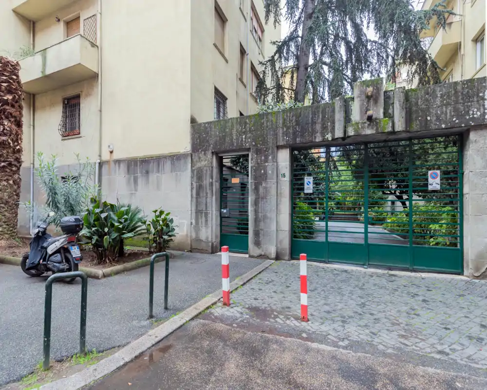 Vista esterna dell’ingresso condominiale con cancello carrabile e porta pedonale, delimitato da muri in cemento e piante.