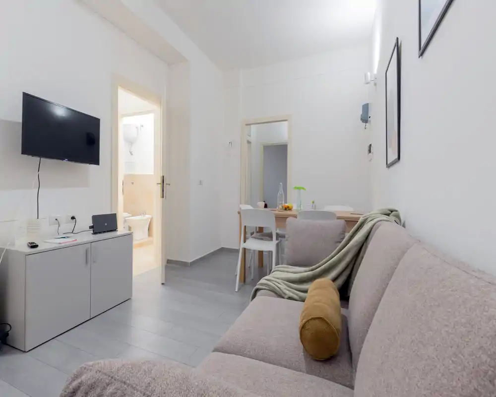 Soggiorno moderno e luminoso con divano confortevole, TV a parete e tavolo da pranzo sullo sfondo — allestimento curato e buona illuminazione ideale per un annuncio.