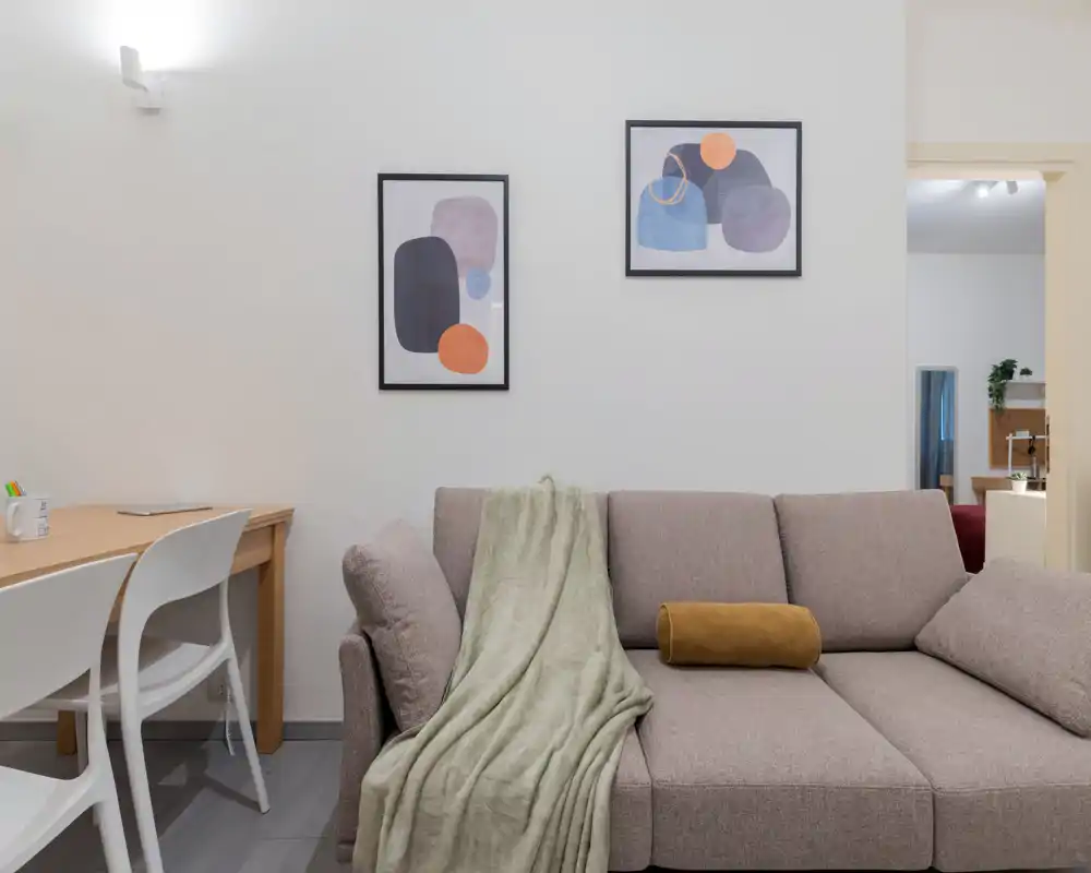 Zona giorno moderna e luminosa con un comodo divano grigio, plaid morbido e cuscini decorativi, affiancata da una piccola scrivania e quadri minimal: un angolo accogliente perfetto per rilassarsi o lavorare.