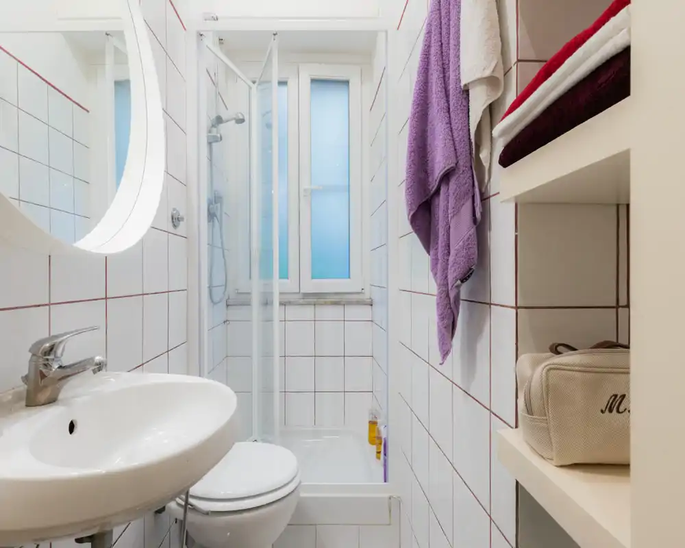 Bagno compatto e ben illuminato con lavabo, WC e box doccia; piastrelle bianche e asciugamani piegati ordinatamente su scaffali aperti.