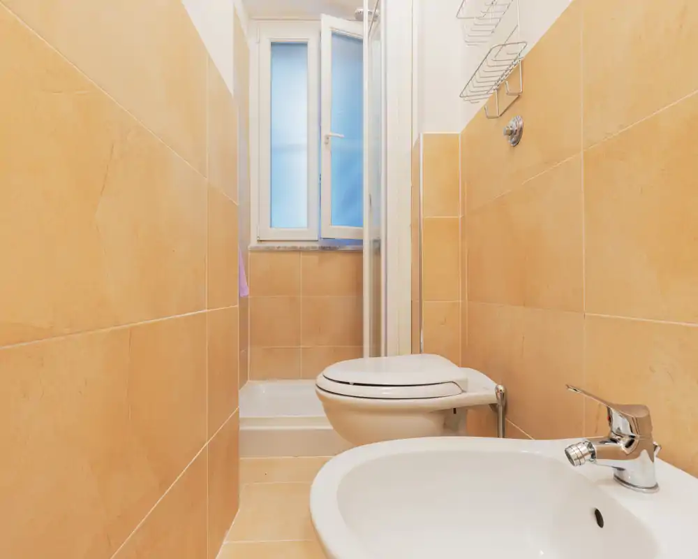 Bagno compatto e ben illuminato con pareti piastrellate beige, wc, bidet e una piccola doccia sul fondo sotto una finestra satinata.