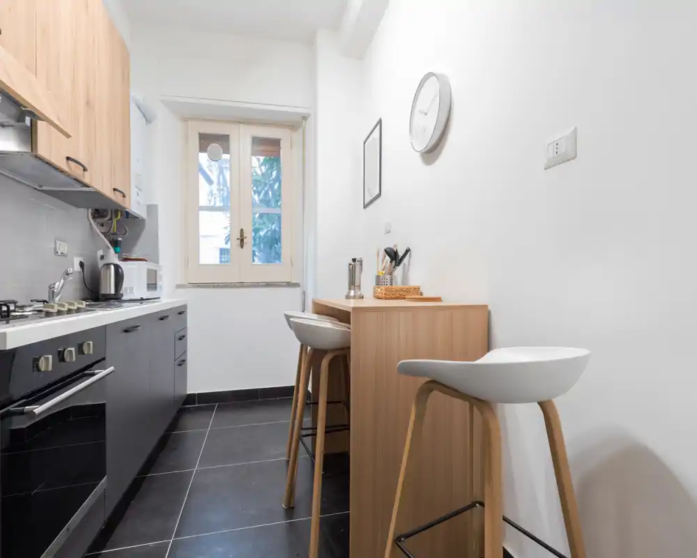 Cucina compatta e moderna con forno integrato, piccoli elettrodomestici sul piano e un piccolo bancone in legno con sgabelli; finestra luminosa e dettagli in legno caldo.