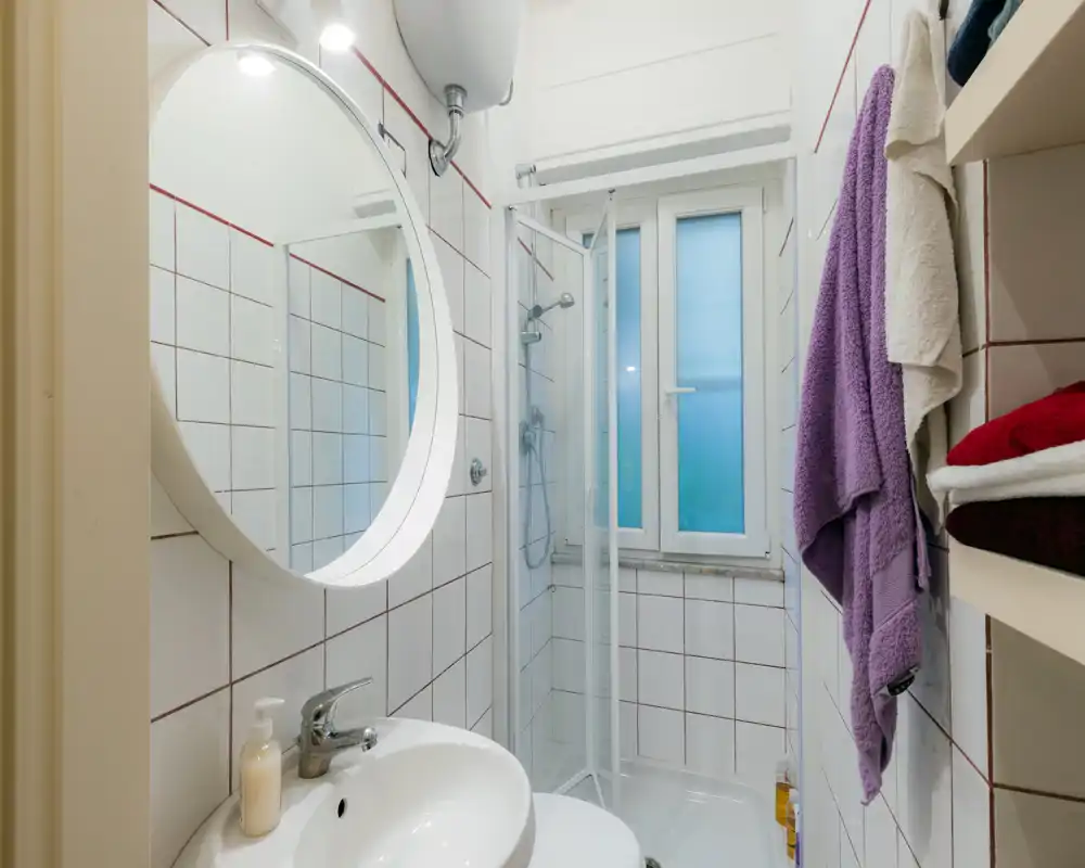 Bagno compatto e ben illuminato con specchio circolare, lavabo e box doccia angolare; pareti piastrellate bianche e scaffalatura per asciugamani lo rendono funzionale.