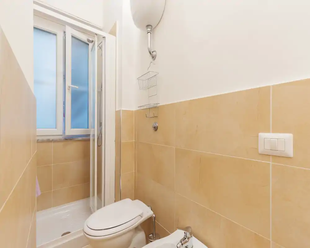 Bagno compatto e ben illuminato con box doccia, WC e bidet; piastrelle beige e finestra satinata per la privacy.