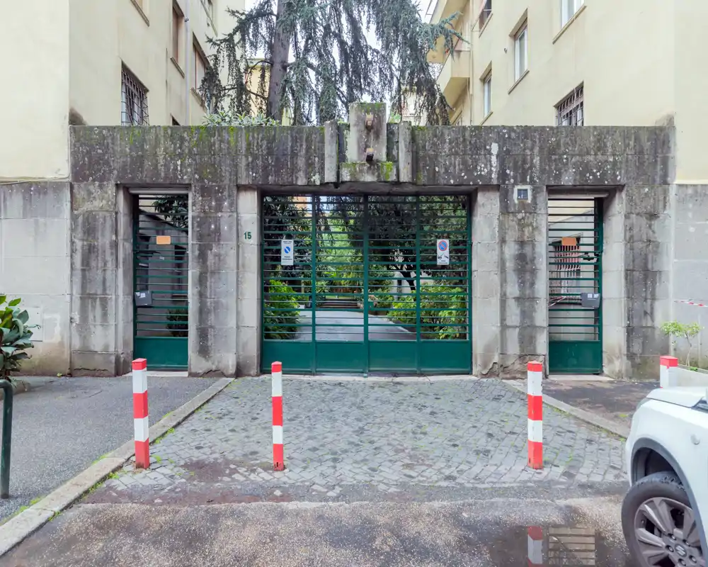 Ingresso esterno recintato di un edificio che conduce a un cortile verde; cancelli in metallo a listelli, facciata in pietra e dissuasori davanti.