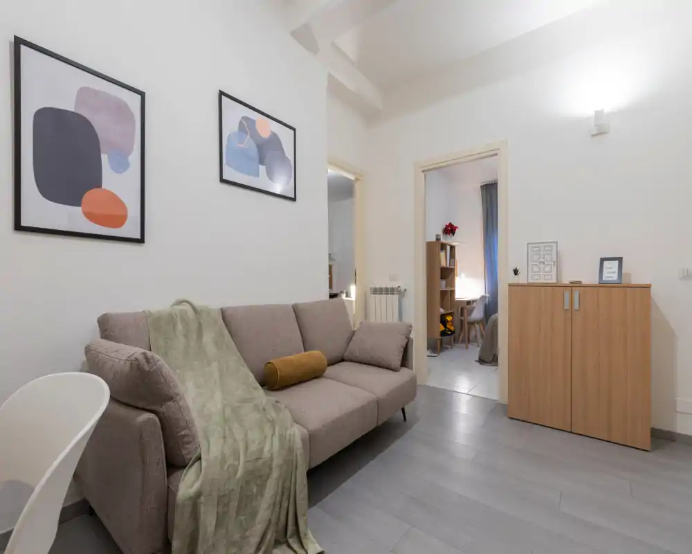 Zona giorno moderna e luminosa con un divano comodo da tre posti, plaid decorativo e quadri alle pareti. Toni neutri e allestimento ordinato creano uno spazio accogliente e tranquillo.