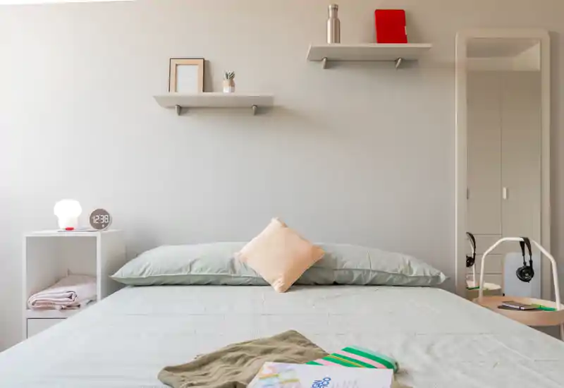 Dormitorio luminoso y de estilo con cama bien hecha, ropa de cama en tonos pastel, cojín decorativo central y estanterías minimalistas sobre la cabecera — acogedor y bien iluminado.