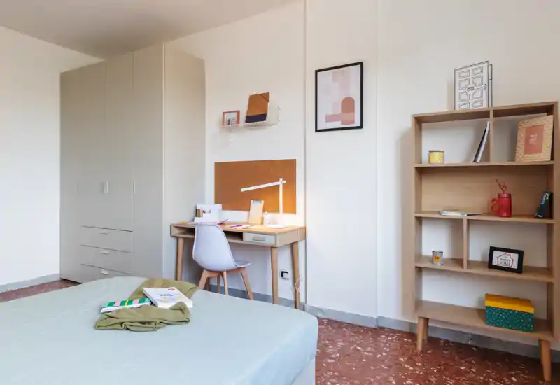 Dormitorio moderno y luminoso con cama bien hecha, armario, escritorio y estantería — ideal para estudiantes o profesionales que buscan una habitación ordenada.